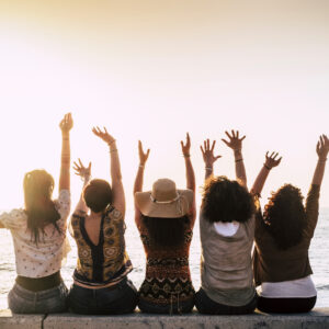 Group of happy and excited females friends enjoy beach day and friendship all together sitting agains the ocean moving raised up arms and dancing. Travel and summer holiday destination happy lifestyle